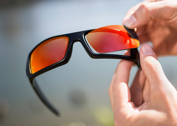 A close-up of hands swapping the orange lenses on black framed sunglasses.