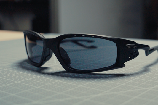 Black Oakley Plantaris sunglasses with blue-tinted lenses on a gray grid cutting mat.