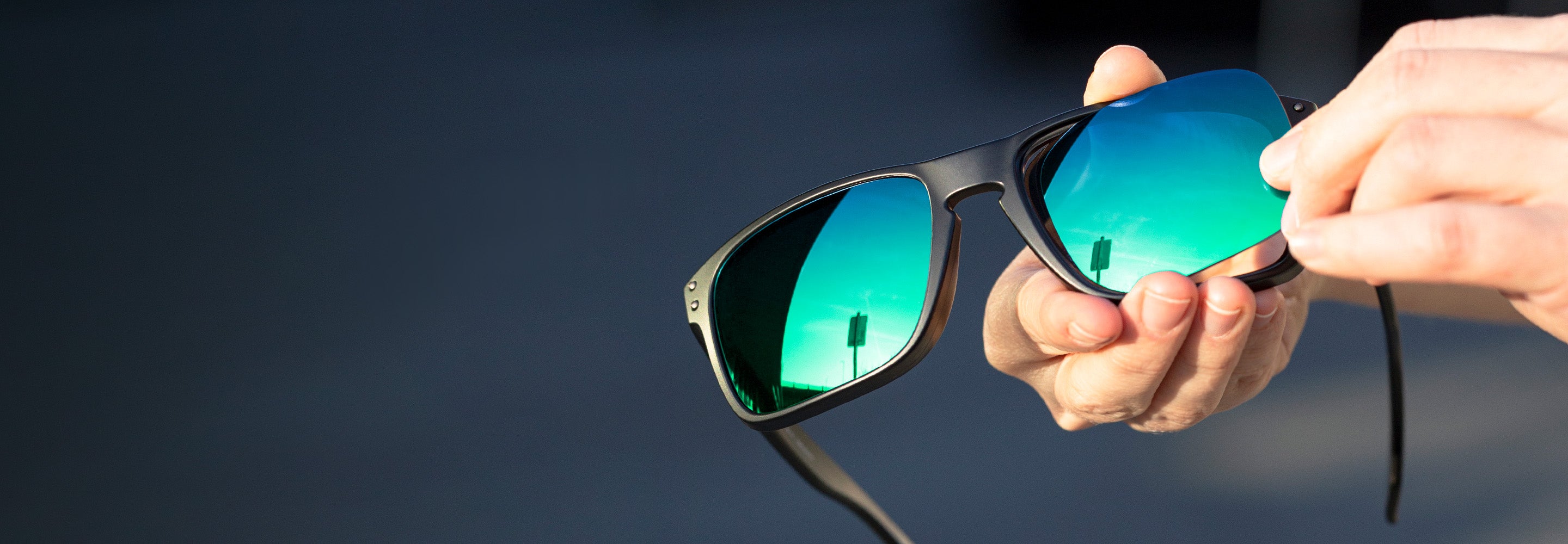 Close-up of a person's hands holding a pair of Oakley Holbrook black sunglasses with vibrant green reflective lenses. 