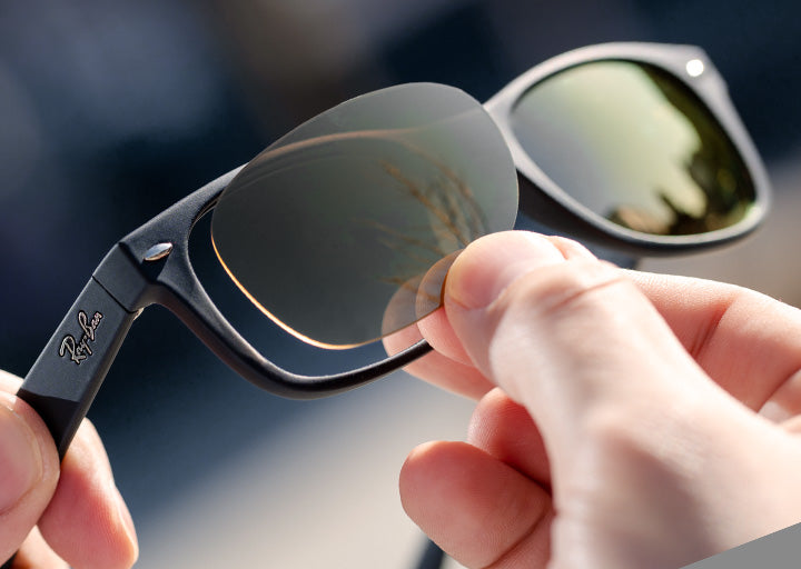 Close-up of hands holding a pair of Ray-Ban sunglasses with reflective green lenses.