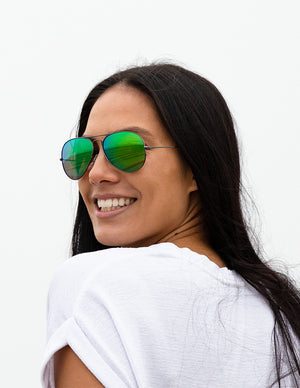 Shoulders-up view of a model with long, dark hair smiling and wearing sunglasses with Emerald Green Lenses.