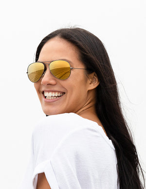 Shoulders-up view of a model with long, dark hair smiling and wearing sunglasses with Flare Gold Lenses