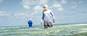 Two fishermen in hats and sunshield face wraps standing in thigh-deep water. One is closer to the camera, one is facing away in the background casting a fishing rod.