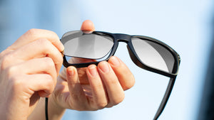 Hands holding and installing a grey lens on black sunglass frames, against a blue backdrop.