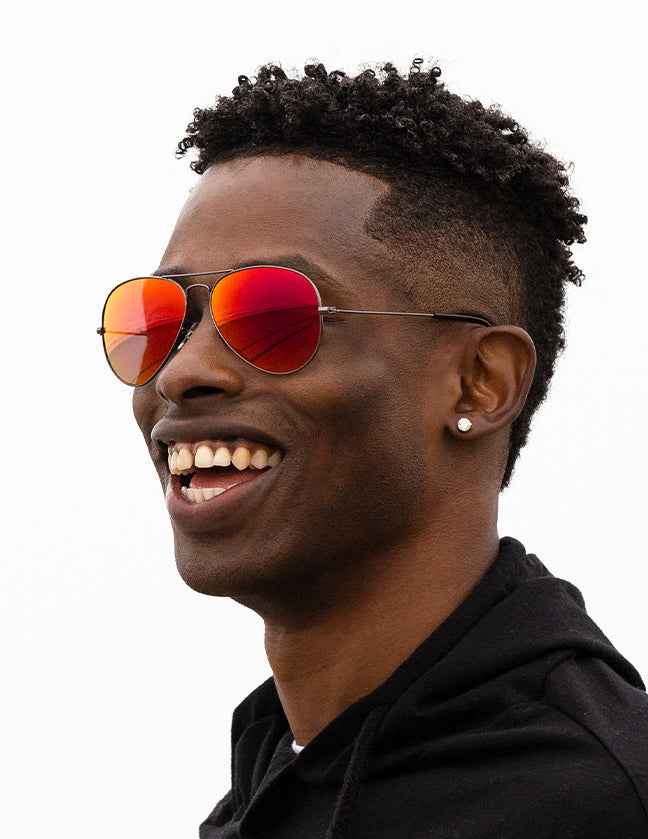 Shoulders-up view of a model with short, dark hair smiling and wearing sunglasses with dark red midnight sun lenses