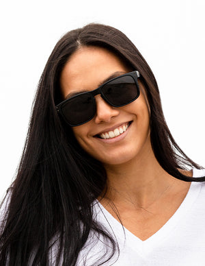 Shoulders-up view of a model with long, dark hair smiling and wearing sunglasses with Stealth Black lenses.