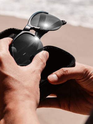 Hands holding a pair of black sunglasses with black lenses against a sand and beach backdrop.