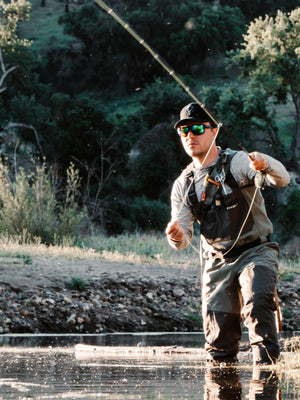 A fisherman in waders standing in a creek, fly fishing in sunglasses with Emerald Green lenses.